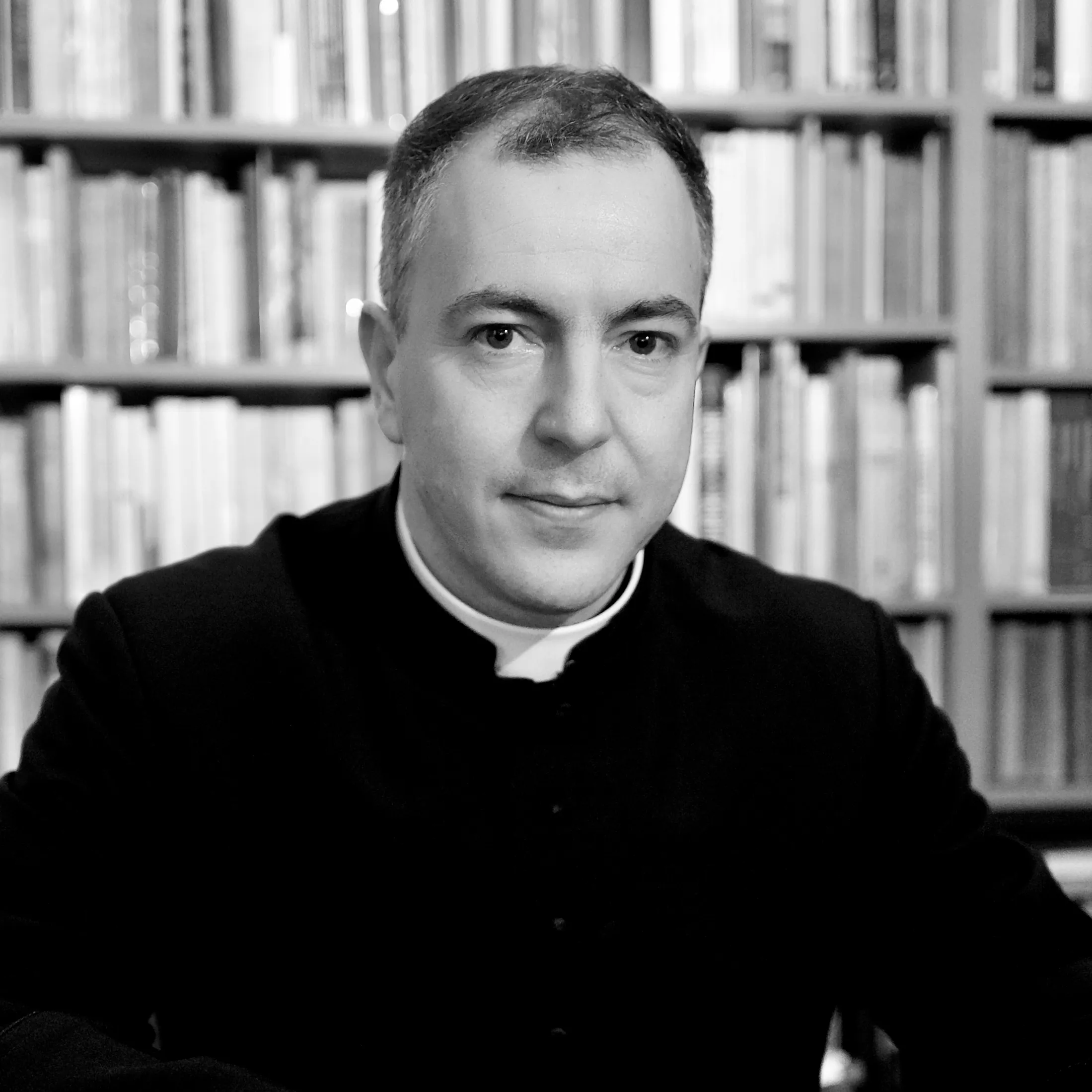 Portret alb-negru al preotului care însoțește activitățile asociației, în fața unei biblioteci.